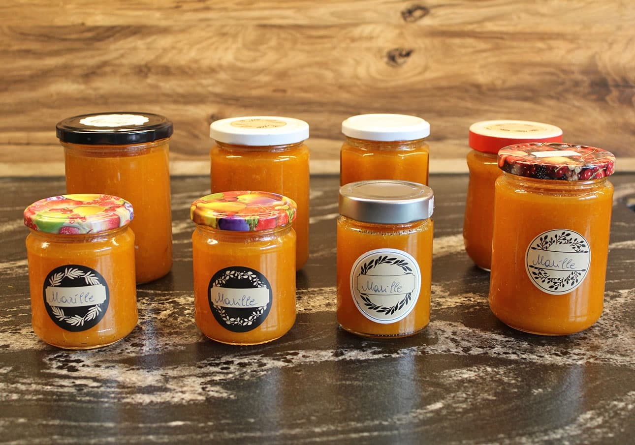 Seven jars of orange marmalade with varied labels and lids on a dark countertop