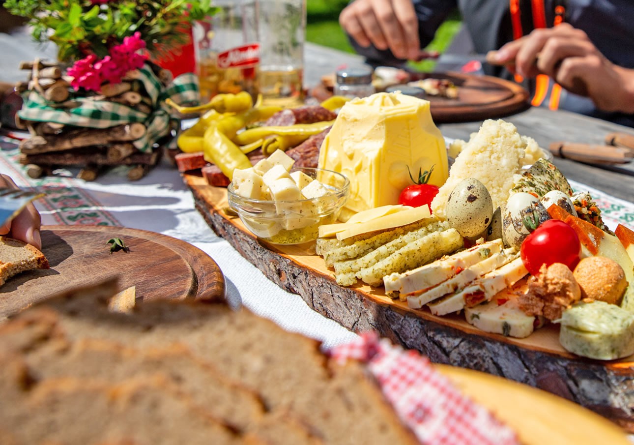 A wooden platter with assorted cheeses, bread, and vegetables on an outdoor table
