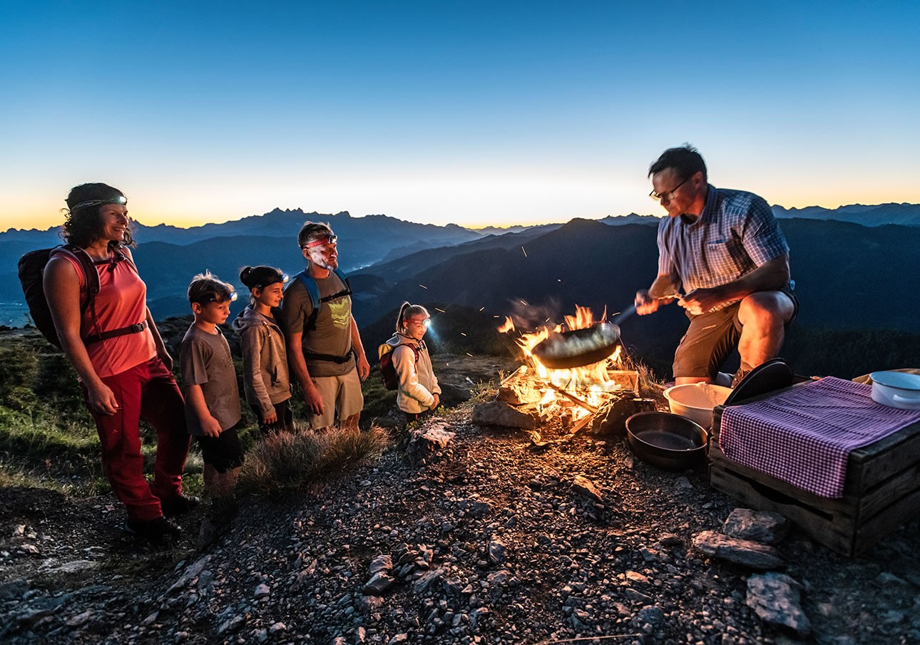 A group watches a man cook over a campfire on a mountain at sunset