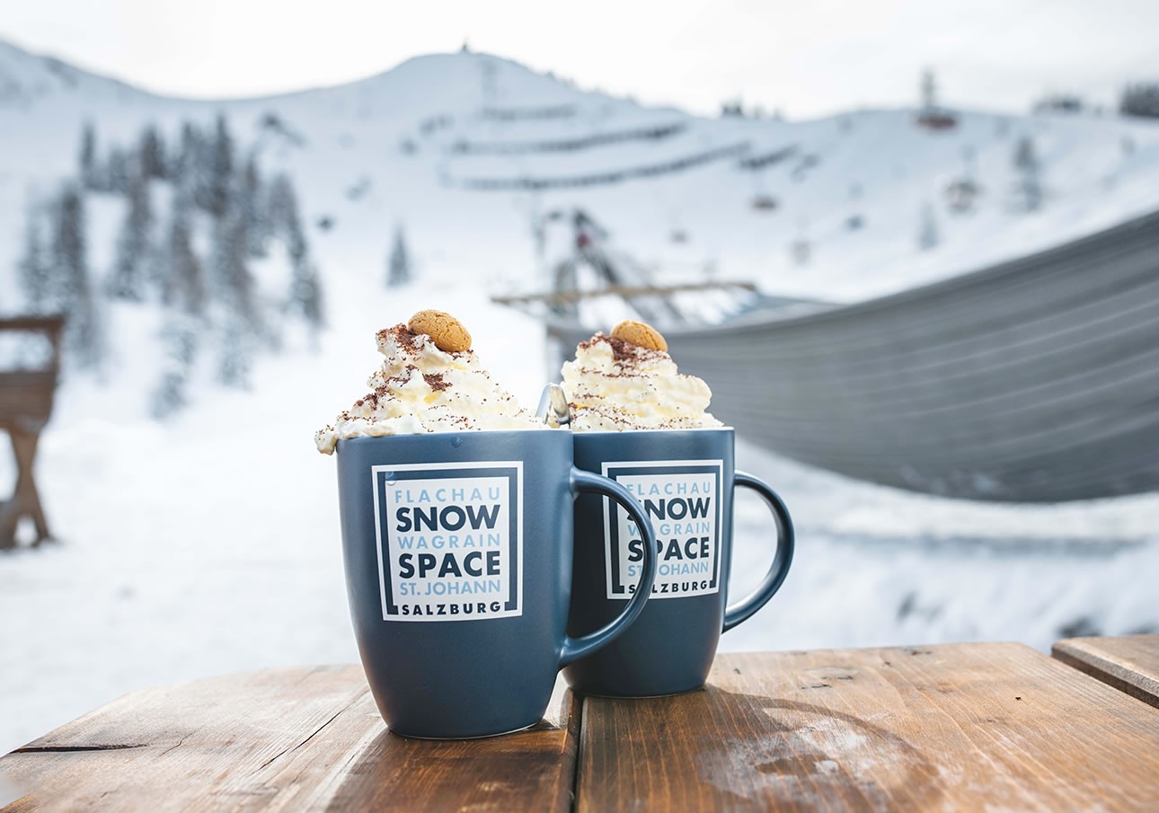 Two blue mugs of whipped cream-topped drinks on a snowy mountain patio table