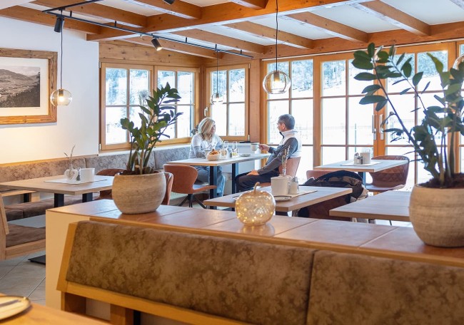 Two people sit at a table in a cozy cafe with large windows and potted plants
