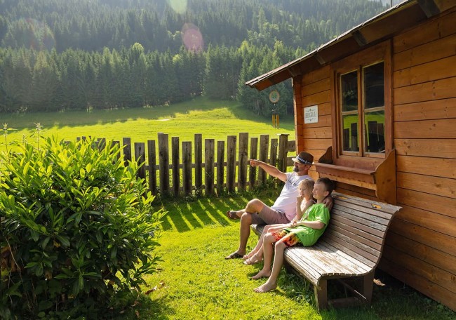 A man and two children relax on a bench by a wooden cabin, overlooking a grassy field