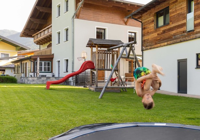 Child doing a flip on a trampoline in a backyard with a playground and houses nearby
